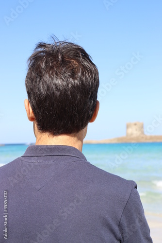 Dark-haired man on his back in blue T-shirt looking at the sea, contemplating the Torre Pelosa, in Costa Smeralda, Sardinia.
