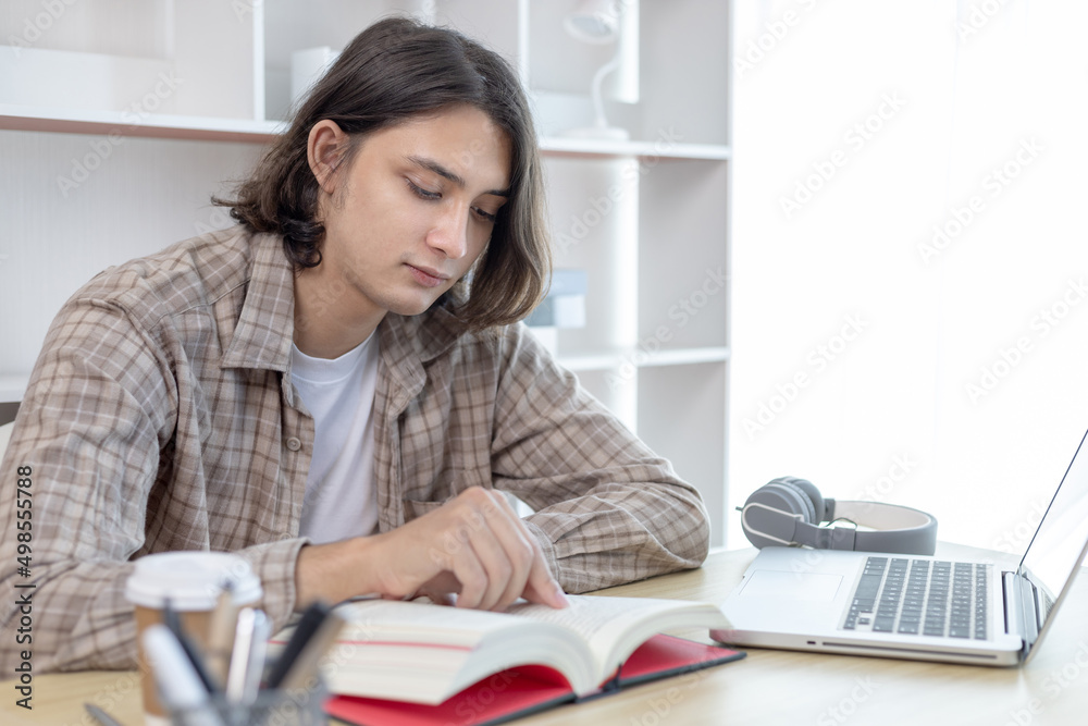 Young male student reading a book in a private office, Long distance education, Textbooks from books, Find out more, In the office there is a computer or laptop placed on the table, Study. Young male student reading a book in a private office, Long distance education, Textbooks from books, Find out more, In the office there is a computer or laptop placed on the table, Study.