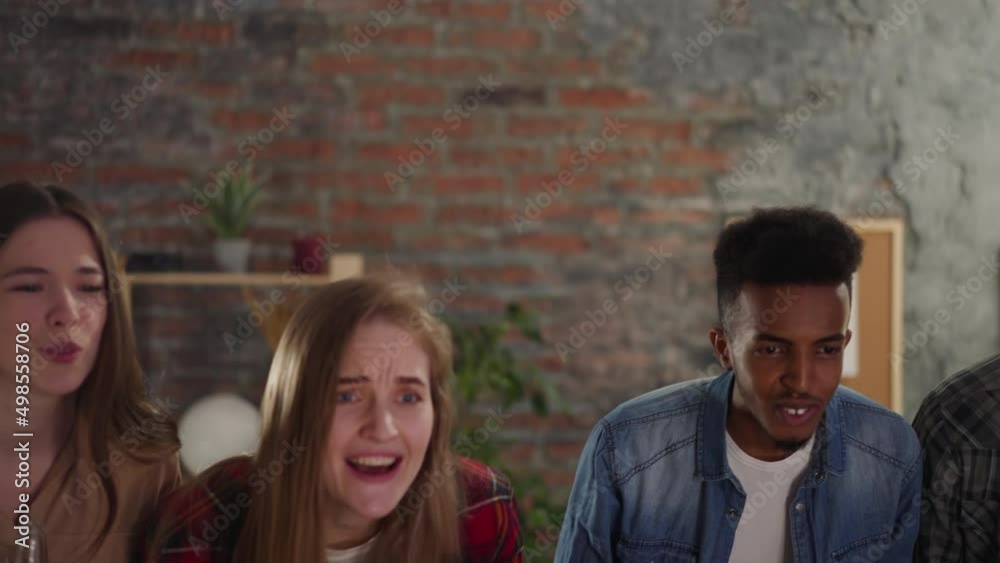 Excited young women hug and drink beer watching football game with black guys in apartment slow motion. Happy sports fans multinational group at championship Excited young women hug and drink beer watching football game with black guys in apartment slow motion. Happy sports fans multinational group at championship