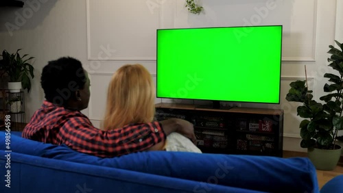 Green screen TV set multiethnic family couple in love embrace watching movie series on TV. Back view husband and wife looking at TV chroma key mock up display. Man and woman sitting on couch at home