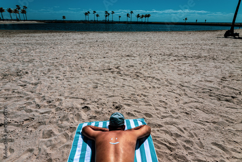 The guy lies on his stomach on a beach towel, a smiley face is drawn with cream on his back. Soft focus