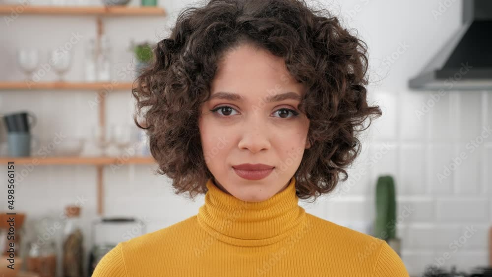 Close up face hispanic curly woman looking at the camera standing at home kitchen. Headshot beautiful female student freelancer in a yellow turtleneck