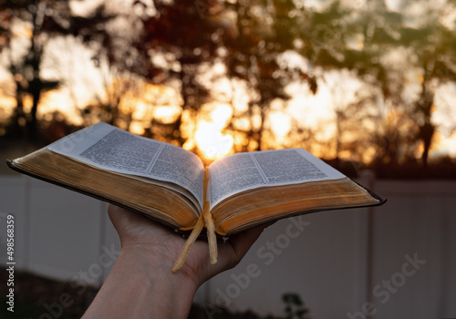 Close up woman outstretching hand with open Bible near bright sunset in the background. copy space.