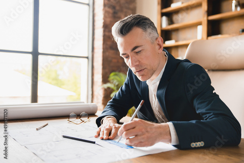 Male mature middle-aged engineer architect drawing blueprints, working on architectural plans in office sitting at the desk, construction industry concept.