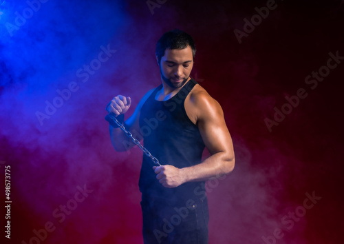 muscular man with a chain. sportsman posing in black t-shirt, jeans. studio portrait. red background blue smoke around the guy