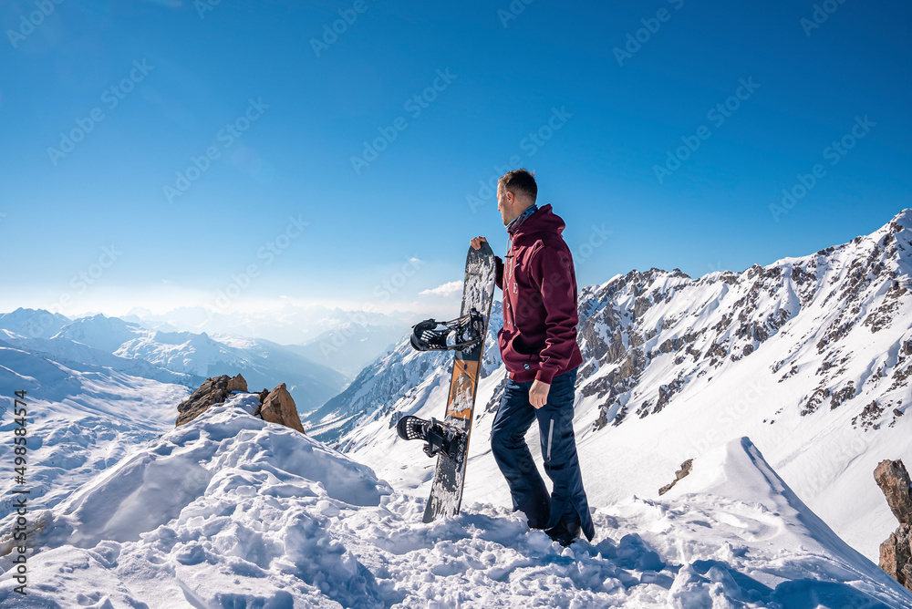 St. Anton am Arlberg. March 10, 2022. Young man holding snowboard while standing on top of snowy mountain, Snowboarder stands on the background of beautiful view of snowcapped mountains