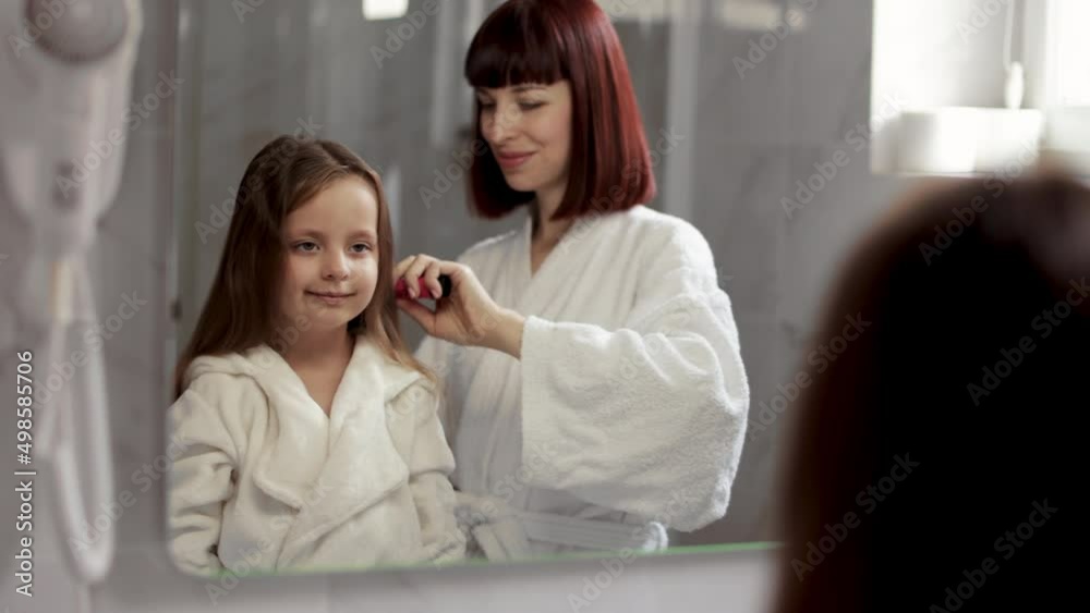 Beautiful young mother In white bathrobe combing hair of her little daughter after taking bath. Loving mom brushing hair of cute little girl standing in front of a mirror in the bathroom. Beautiful young mother In white bathrobe combing hair of her little daughter after taking bath. Loving mom brushing hair of cute little girl standing in front of a mirror in the bathroom.