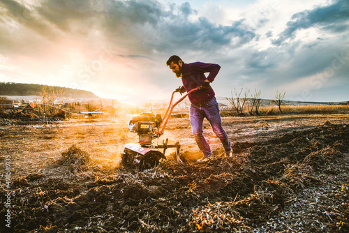 organic farming man cultivates the ground at sunset with a tiller  preparing the soil for sowing photo