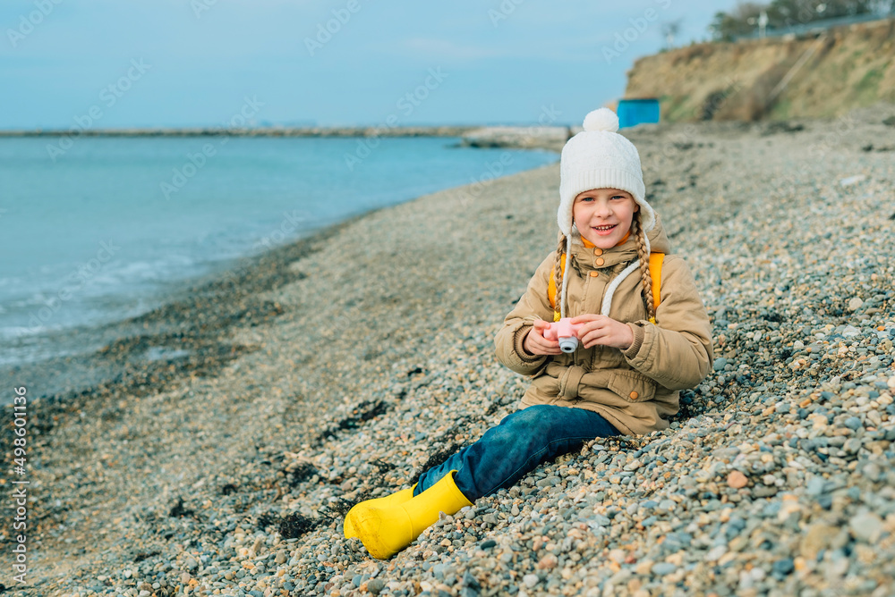 little girl sits in autumn, spring on the beach, looks at the camera with a smile, holds a children's pink camera in her hands. the child is passionate about his hobby: photography little girl sits in autumn, spring on the beach, looks at the camera with a smile, holds a children's pink camera in her hands. the child is passionate about his hobby: photography