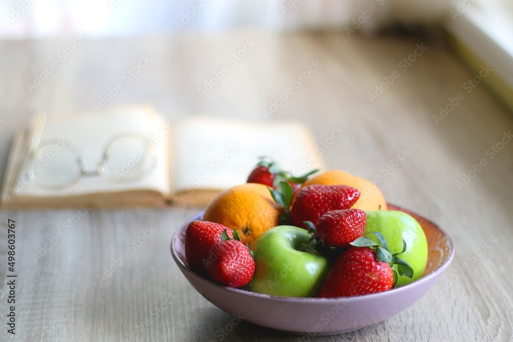 Bowl of apples, oranges and strawberries, open book and reading glasses on the table. Selective focus. Bowl of apples, oranges and strawberries, open book and reading glasses on the table. Selective focus.