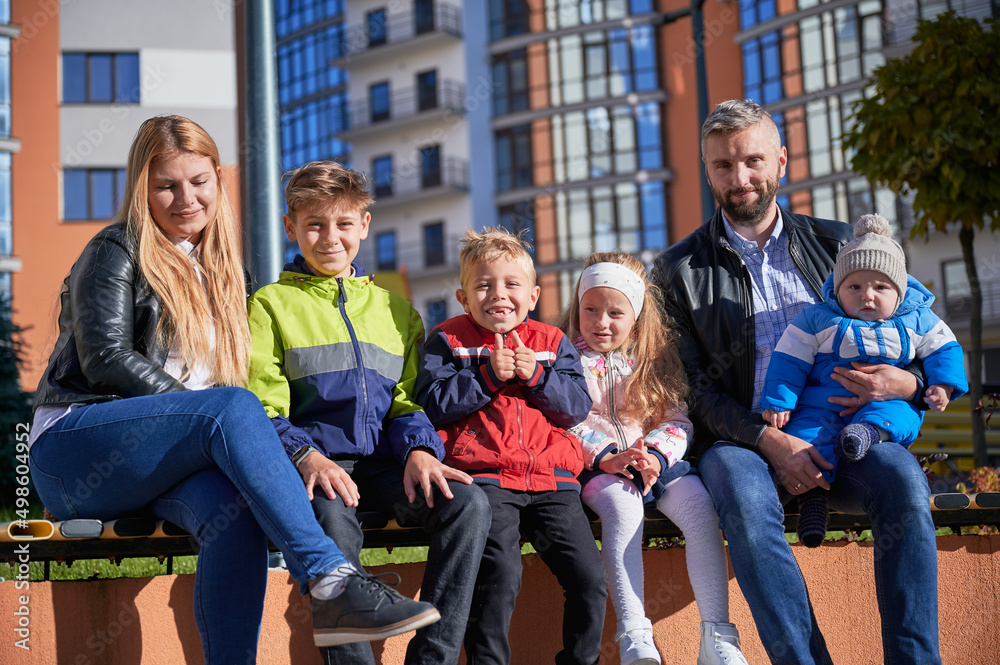 Obraz premium Portrait of happy family - father, mother and children having fun together on playground. Parents and kids sitting on bench, looking to camera. Modern residential buildings on background.
