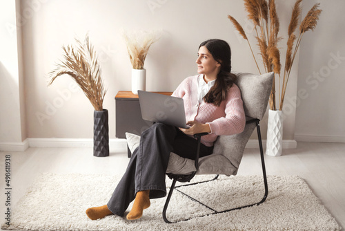 Full body beautiful confident woman 30 years old working remote on laptop from cozy home sitting in comfortable chair with trendy interior photo