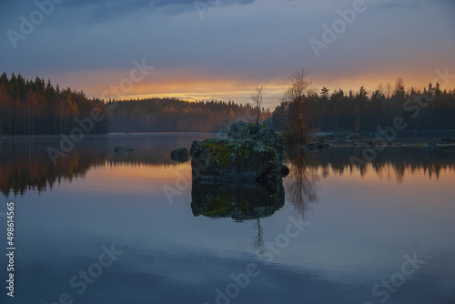 Sunrise on a small calm Finnish lake with a large stone and trees in the middle, blue and orange sky above blue water © Olga Begak Art