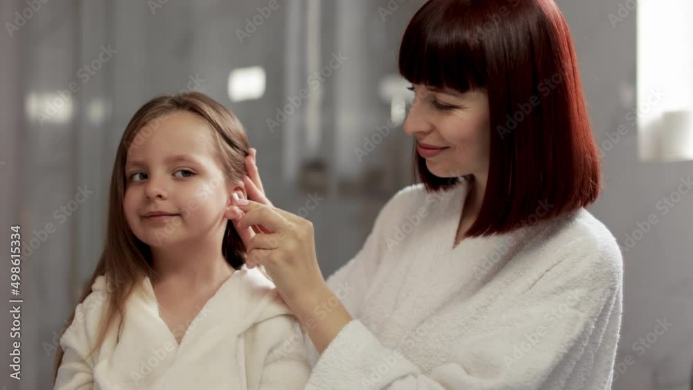 Close up of small blond haired cute girl in white bathrobe after bath, standing in bathroom at home, while her affectionate mother uses cotton swabs and cleans the ears. Mom and little girl at home. Close up of small blond haired cute girl in white bathrobe after bath, standing in bathroom at home, while her affectionate mother uses cotton swabs and cleans the ears. Mom and little girl at home.