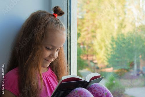 a sweet, dark-skinned little girl in a pink T-shirt and shorts sits at home alone in quarantine on the window sill in the summer and reads a book with interest. Lok Down education. copy space