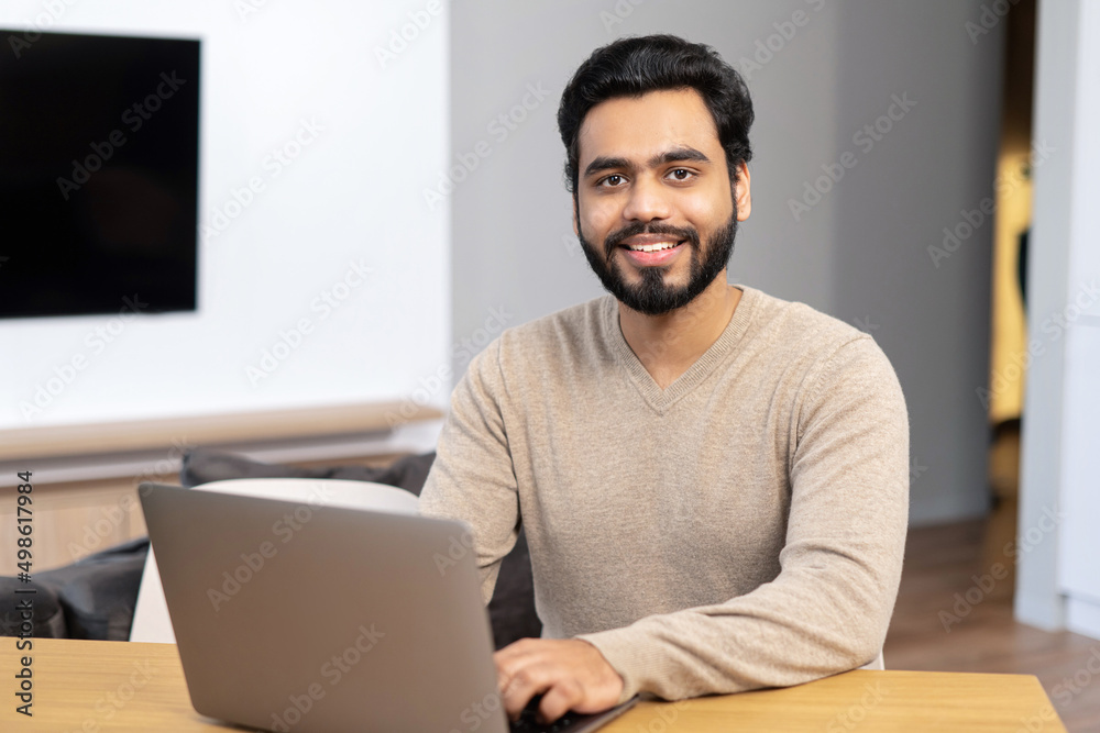 Cheerful Indian man wearing casual wear sitting at the table and using laptop at modern home office, glad ethnic latin male entrepreneur typing on keyboard Cheerful Indian man wearing casual wear sitting at the table and using laptop at modern home office, glad ethnic latin male entrepreneur typing on keyboard