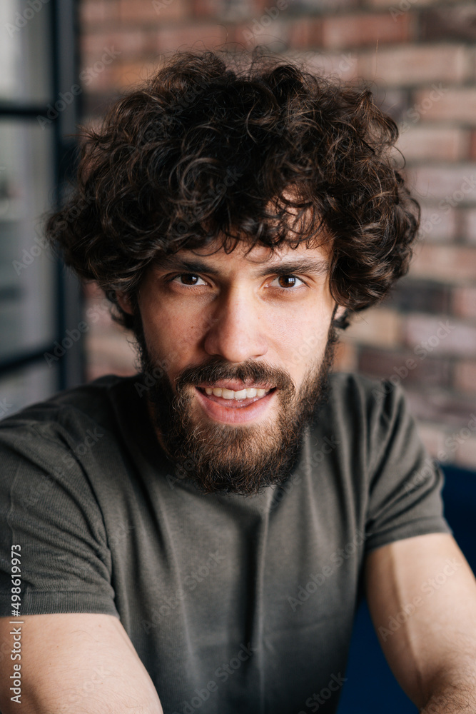 Close-up vertical portrait of handsome young man looking at camera sitting on chair in cozy living room with brick wall. Front view of confident curly male posing at home. Close-up vertical portrait of handsome young man looking at camera sitting on chair in cozy living room with brick wall. Front view of confident curly male posing at home.