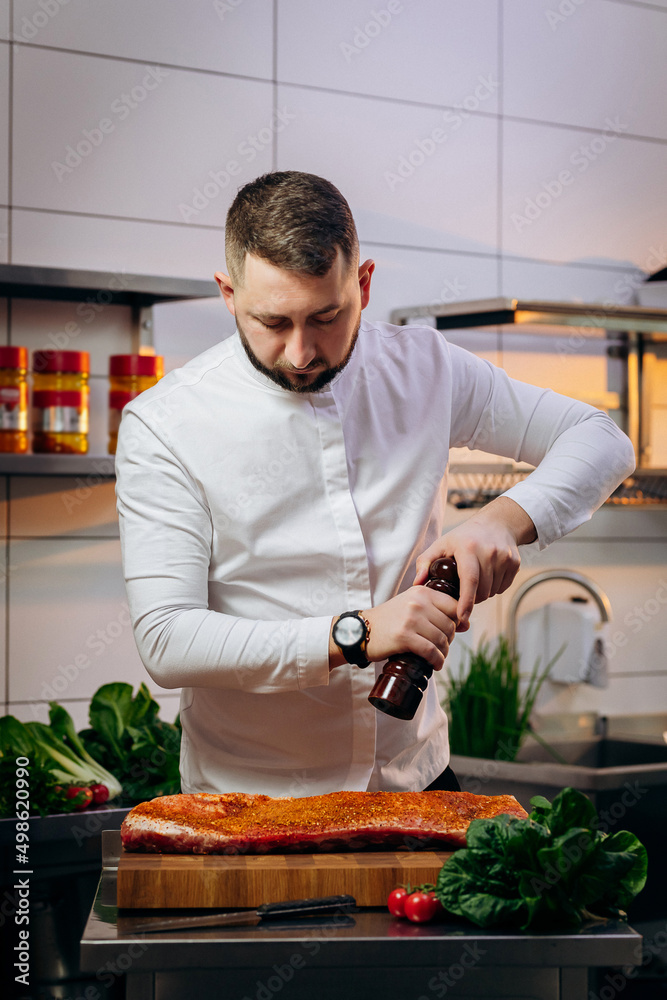 Portrait of handsome chef cooking raw meat in the kitchen. Cook salt and pepper the steak. Concept of Cooking process and preparing dishes. Portrait of handsome chef cooking raw meat in the kitchen. Cook salt and pepper the steak. Concept of Cooking process and preparing dishes.