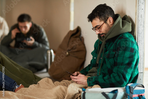 Side view portrait of young man hiding in shelter during war or crisis and holding smartphone photo