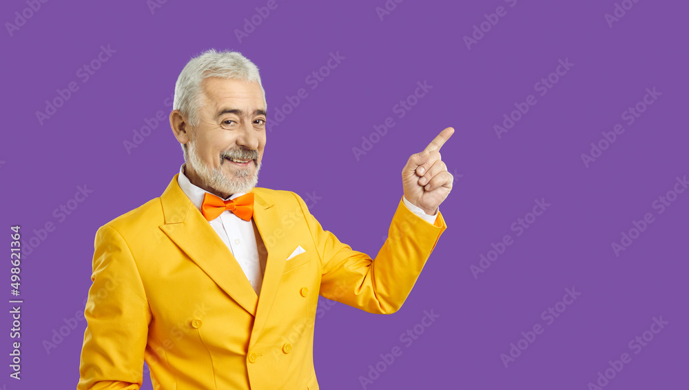 Happy eccentric handsome bearded senior man in bright yellow suit and bow tie isolated on solid purple background smiles and shows something, pointing his index finger at copy space on the side Happy eccentric handsome bearded senior man in bright yellow suit and bow tie isolated on solid purple background smiles and shows something, pointing his index finger at copy space on the side