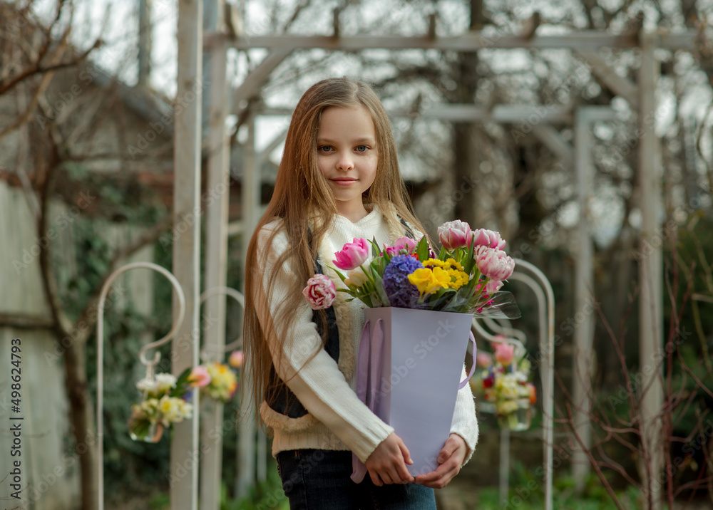 Cute smiling little girl with bouquet of spring flowers in hands. Portrait of kid with long hair with with seasonal floral gift on rural background. Concept of Easter or Mother Day celebration