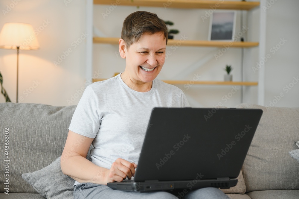 Technology, people and communication concept - Happy senior woman with tablet pc computer having video chat at home. Modern middle-aged 50s grandmother sit relax on couch in living room using laptop.