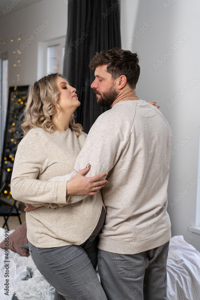 Fototapeta premium Happy family concept. Husband hug belly pregnant wife standing indoor living room near sofa