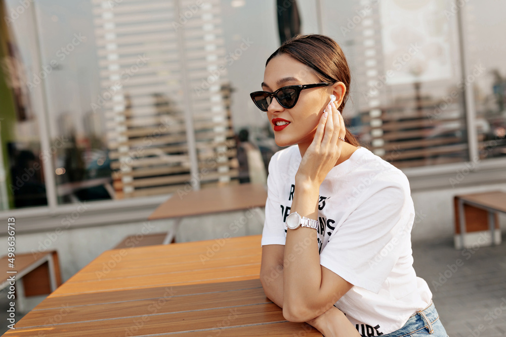 Fashionable lovely woman with dark hair and red lips is wearing glasses and touching headphones while sitting in sunlight Fashionable lovely woman with dark hair and red lips is wearing glasses and touching headphones while sitting in sunlight