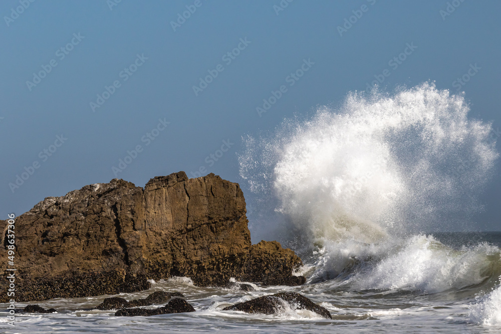 Large wave breaks on offshore rock near Malibu, California, sending ball of spray into the air, curling backwards. Another giant rock nearby. 
