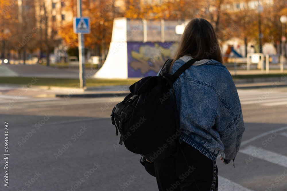 Fototapeta premium Rear view of unrecognizable teenage girl with backpack walking on street. A Young woman walks down city street, black backpack on his back.