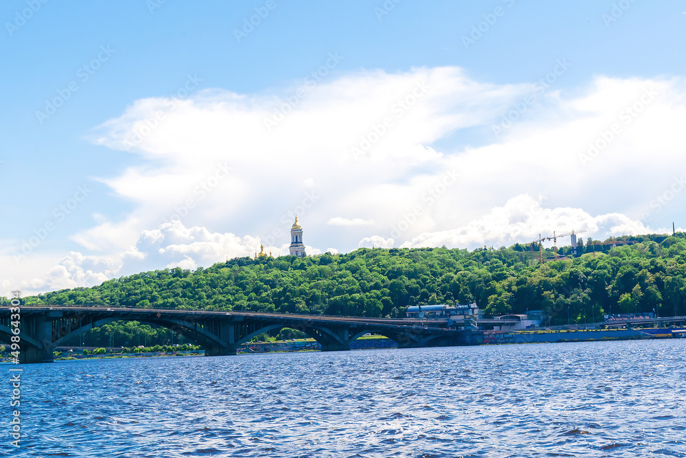 Scenic nature travel cityscape river Dnipro with blue water and bridge at sunny summer day in Kiev. View from inside river of green bank of city park with trees and Lavra church temple