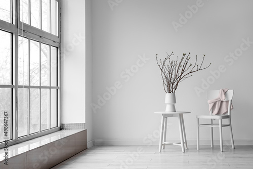 Fotografia Vase with tree branches on table and chair with sweater near white wall in room