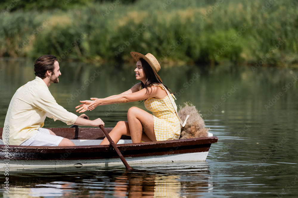 smiling woman in straw hat sitting out outstretched hands near man in boat.