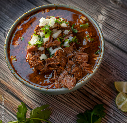 A bowl of BIRRIA or BARBACOA, a traditional Mexican dish with lemons and cilantro on the side over a wooden table, Top view square shot