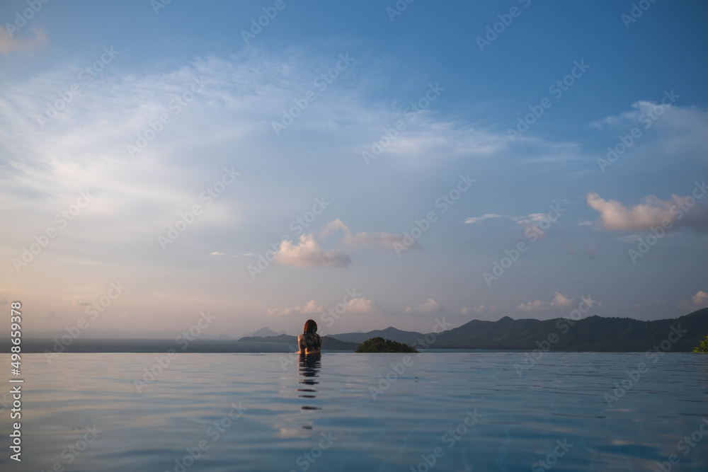 Rear view of a young woman relaxing in infinity swimming pool and looking at a beautiful sunset and the sea view