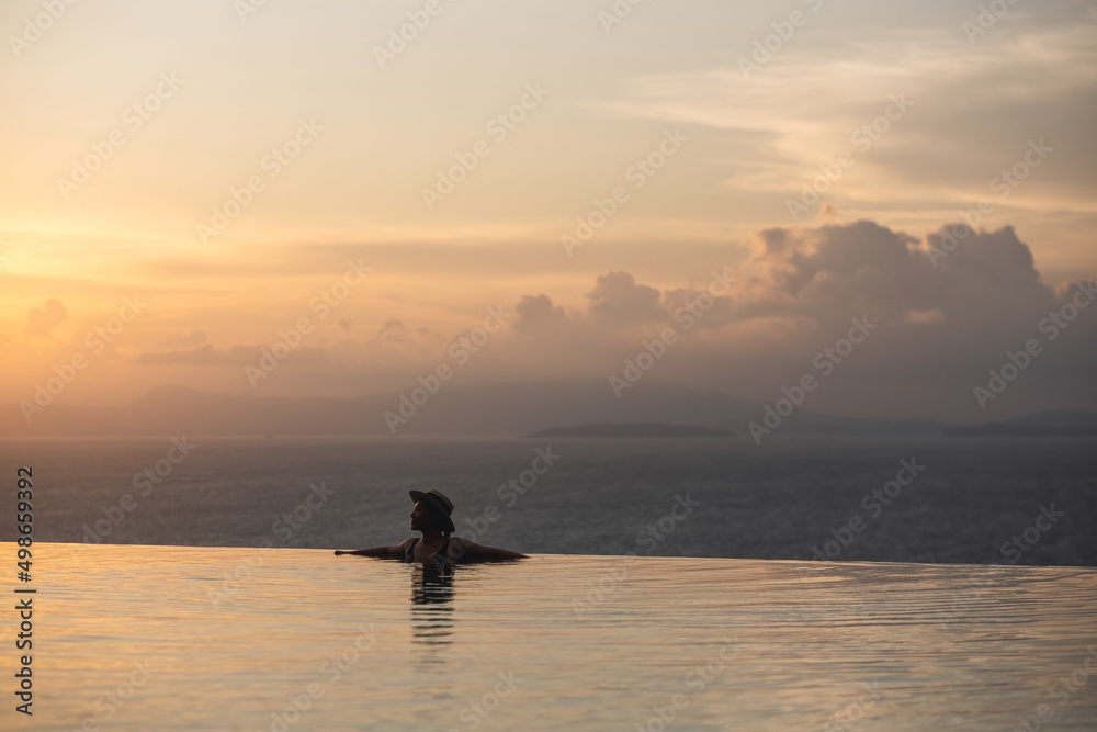 Silhouette of a young woman relaxing in infinity swimming pool and looking at a beautiful sunset and the sea view