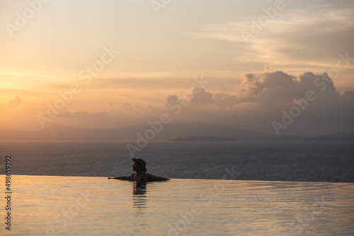 Silhouette of a young woman relaxing in infinity swimming pool and looking at a beautiful sunset and the sea view © Farknot Architect