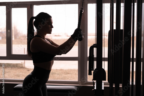 Woman training triceps. A young attractive woman with a perfect body is doing exercises on a cable trainer in the gym silhouette against a window background.