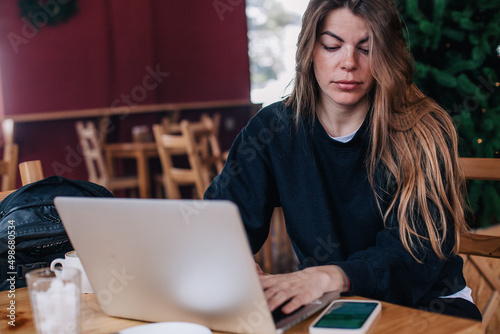 A young beautiful girl in a black jumper in a cafe works with a laptop and a phone