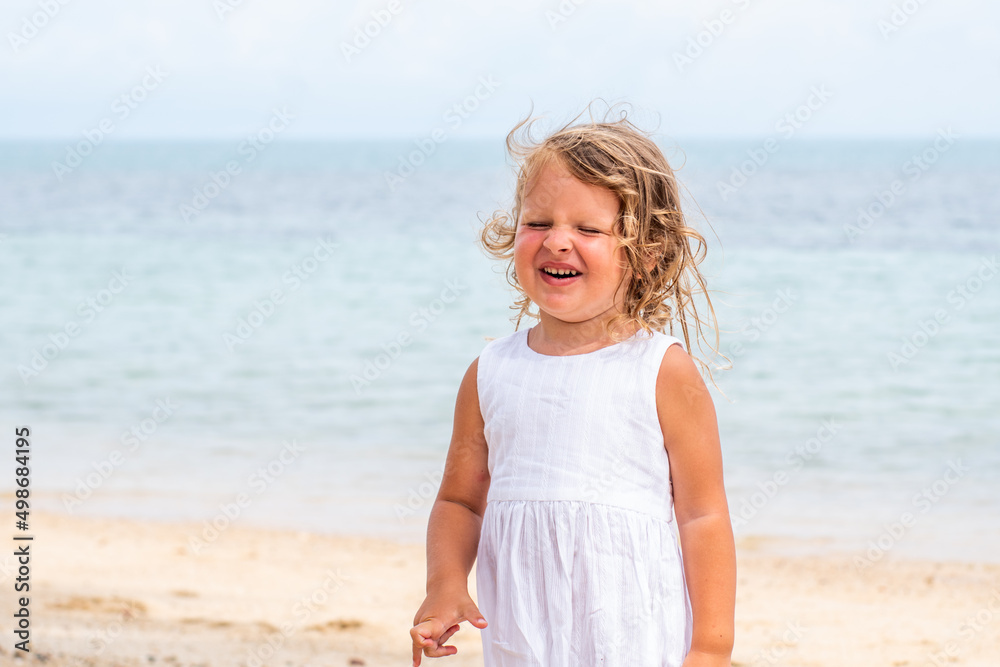 A little girl with curly hair in a white dress walks along a sandy beach on the shore of the sea, ocean and smiles. Sea holidays, travel and beach holidays with children A little girl with curly hair in a white dress walks along a sandy beach on the shore of the sea, ocean and smiles. Sea holidays, travel and beach holidays with children