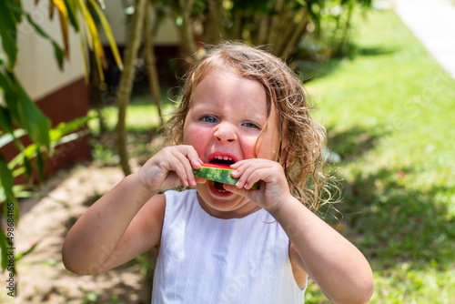 A little cute girl with curly hair has a watermelon. A child eats fruit in the summer in the yard. Healthy eating