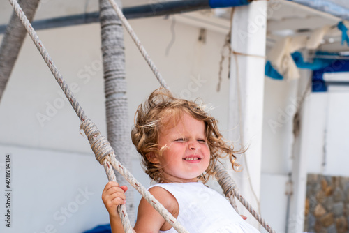 A little girl with curly hair in a white dress is swinging on a swing on the seashore. Cute Child on sandy playing and laughing