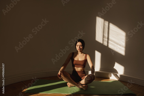 Young caucasian girl practicing yoga, sitting on mat in relaxed pose in morning. Brunette woman exercising at home wearing top and leggings. Wellbeing, mental health care concept