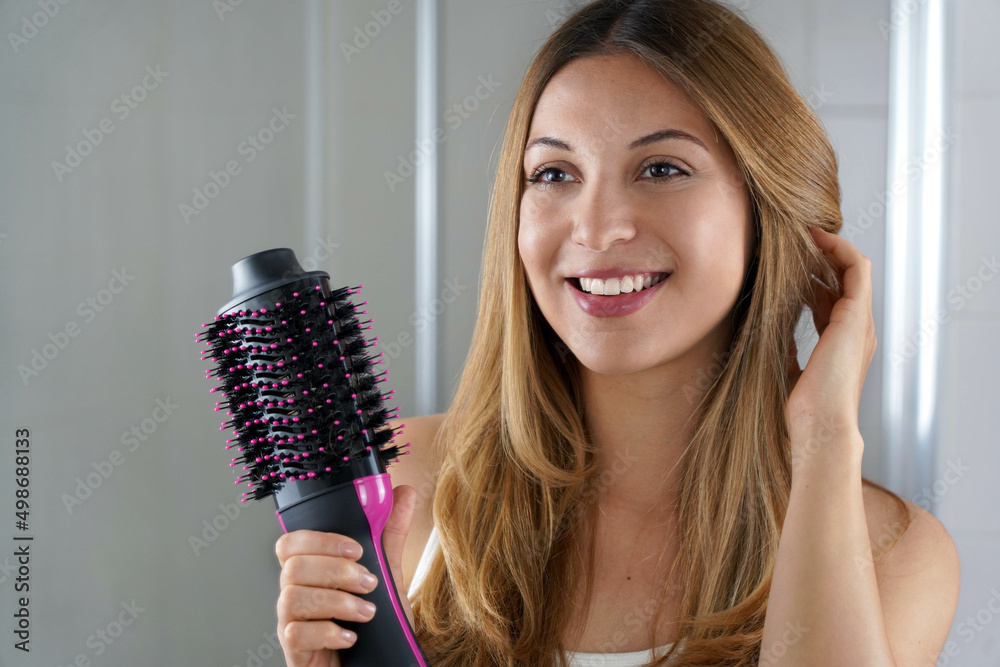 Pleased girl holds round brush hair dryer to style hair in her bathroom at  home. Young woman showing salon one-step hair dryer and volumizer. Stock  Photo | Adobe Stock