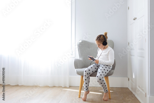 Full body of barefoot girl sitting in armchair and listening to songs from tablet in headphones photo