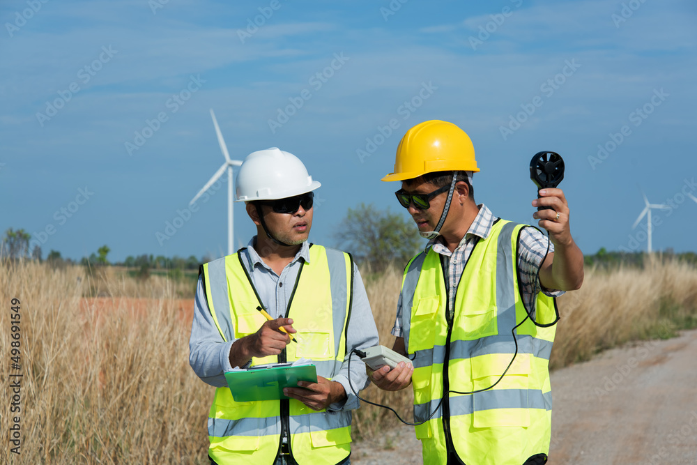 Two maintenance engineer planning discuss Project in wind turbine farm. two person check wind speed measurement Behind is a windmill Two maintenance engineer planning discuss Project in wind turbine farm. two person check wind speed measurement Behind is a windmill