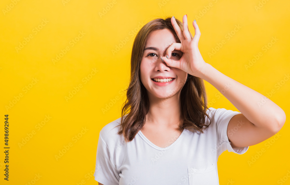 Portrait Asian Thai beautiful young woman standing, She made finger OK symbol sign to agree near two eyes and looking at camera, shoot photo in studio on yellow background, There was copy space Portrait Asian Thai beautiful young woman standing, She made finger OK symbol sign to agree near two eyes and looking at camera, shoot photo in studio on yellow background, There was copy space