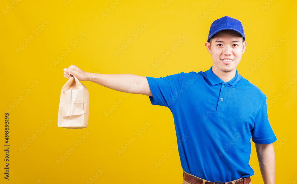 Portrait excited delivery service man smile wearing blue t-shirt and cap uniform hold paper containers for takeaway bag grocery food packet looking to camera, studio shot isolated on yellow background Portrait excited delivery service man smile wearing blue t-shirt and cap uniform hold paper containers for takeaway bag grocery food packet looking to camera, studio shot isolated on yellow background