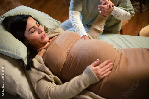 Cropped image of pregnant woman and helpful midwife at home. Woman in casual clothes lying on bed, midwife holding hand. Pregnancy, medicine, home birth concept photo