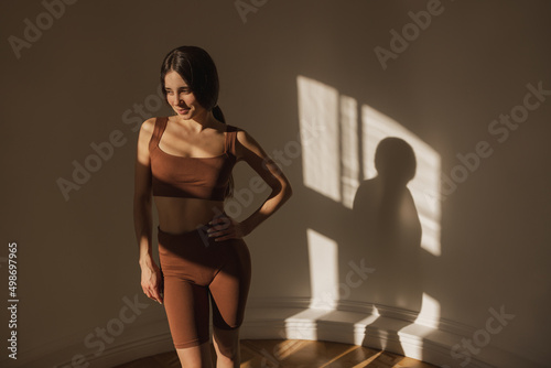 Cute light-skinned young girl keeps her hand on her waist while standing in sunny room with space for text. Brunette wears brown top and leggings. Beauty and health concept